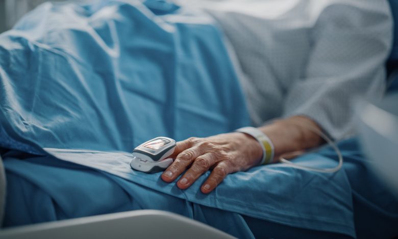 Hospital Ward: Senior Woman Resting in a bed with Finger Heart Rate Monitor / Pulse oximeter showing Pulse. Her Fragile Hands Resting on a Blanket. Focus on the Hand.