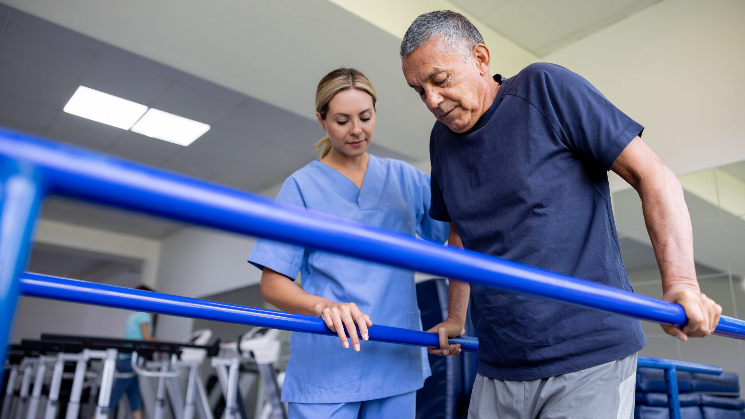 Latin American man doing physical therapy and walking on bars with the assistance of his physical therapist