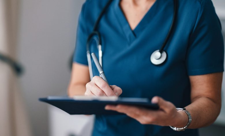 A close-up of a healthcare professional in blue scrubs writing notes on a clipboard, symbolizing care and medical professionalism in a modern medical setting.