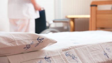 Bed at nursing home with nurse and wheelchair on background.