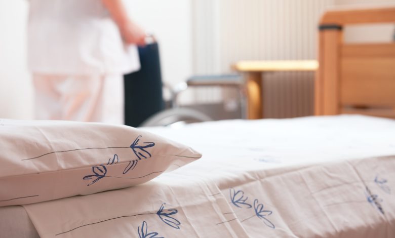 Bed at nursing home with nurse and wheelchair on background.