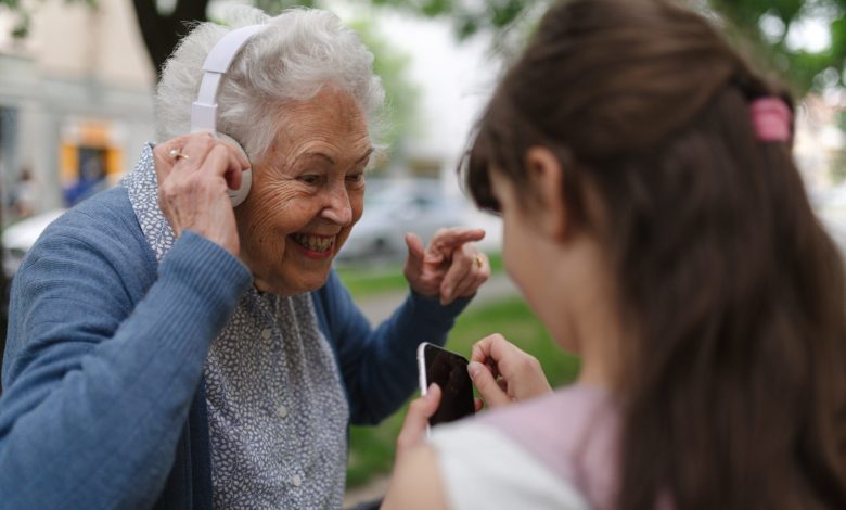 Grandma picking up young girl from school at afternoon. Granddaughter playing music on smartphone to senior grandmother with headphones.