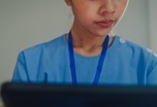 Closeup of young Asian female nurse in blue scrubs sitting on bed while using a digital tablet. Nurse lifestyle concept.