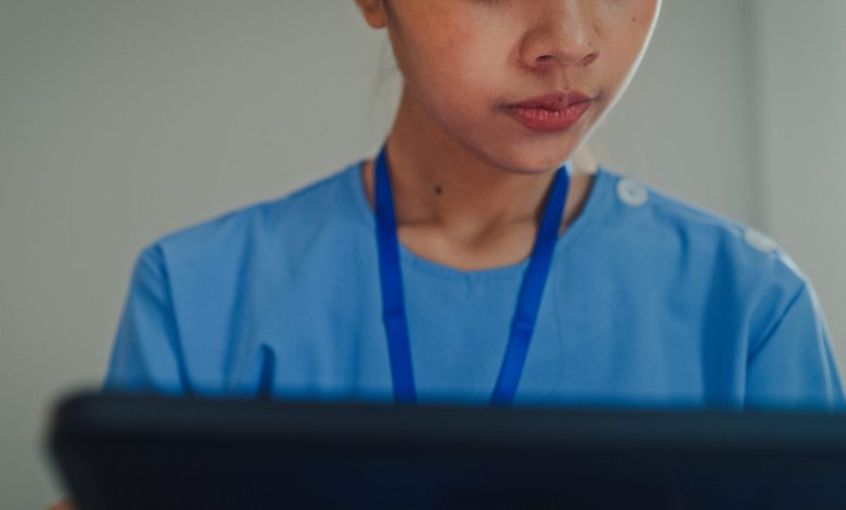 Closeup of young Asian female nurse in blue scrubs sitting on bed while using a digital tablet. Nurse lifestyle concept.