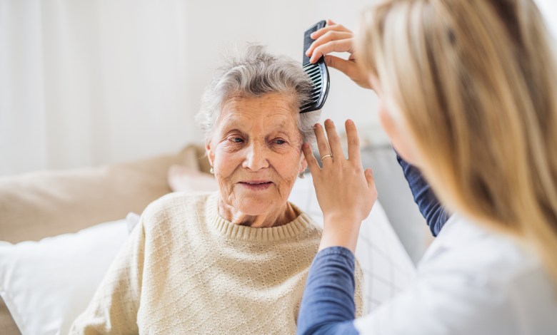 An unrecognizable health visitor combing hair of senior woman sitting on a sofa at home.