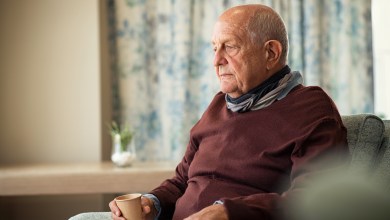Depressed senior man sitting on armchair holding disposable cup of coffee and thinking. Frustrated retired man sitting on sofa and drinking a cup of hot tea. Sad mature man sitting alone at nursing home with sad expression.