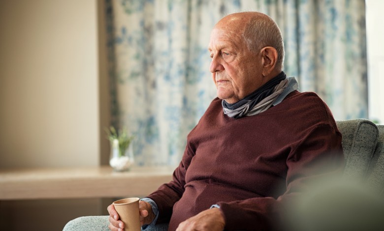 Depressed senior man sitting on armchair holding disposable cup of coffee and thinking. Frustrated retired man sitting on sofa and drinking a cup of hot tea. Sad mature man sitting alone at nursing home with sad expression.